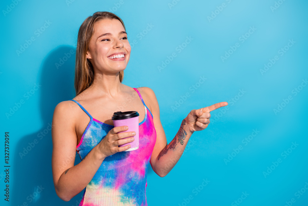 Smiling young woman in colorful casual attire holding a coffee cup and pointing, standing against a bright blue background Smiling young woman in colorful casual attire holding a coffee cup and pointing, standing against a bright blue background