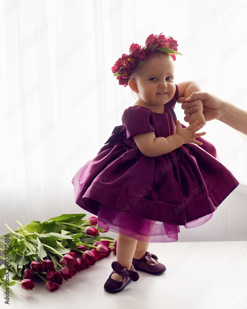 A portrait of a cheerful, beautiful one-year-old girl stands in a burgundy dress at the window of the house. He wears a wreath of flowers on his head