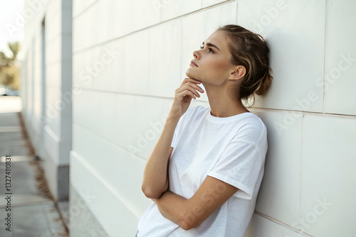 Thoughtful young woman in a stylish white t-shirt poses pensively against a modern urban wall during a sunny day outdoors, capturing a moment of reflection. Free Download Thoughtful young woman in a stylish white t-shirt poses pensively against a modern urban wall during a sunny day outdoors, capturing a moment of reflection. - GFXLoad