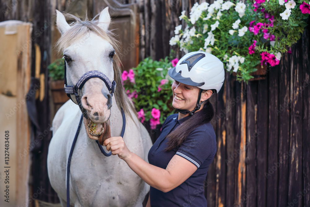 Young woman wearing equestrian helmet standing next to her white Arabian horse, holding animal tongue, smiling. Ranch wooden boxes behind
