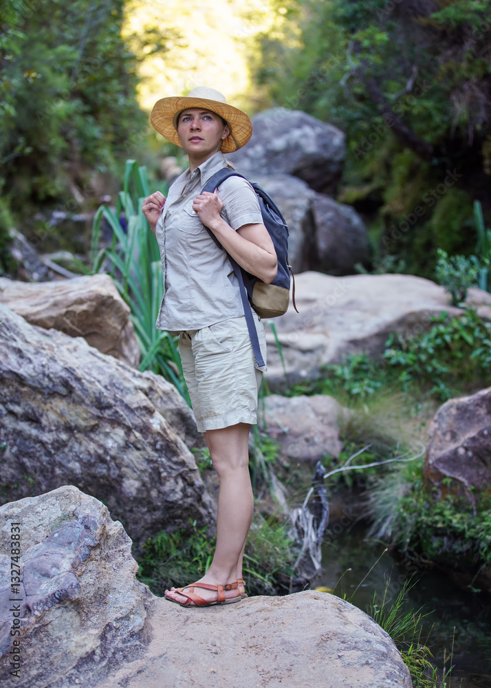 Young woman in beige shirt, shorts and straw haw, backpack on shoulders, hiking rainforest jungle, small creek near