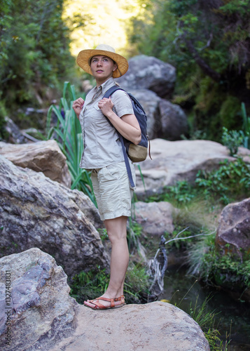 Obraz na płótnie Young woman in beige shirt, shorts and straw haw, backpack on shoulders, hiking rainforest jungle, small creek near w salonie