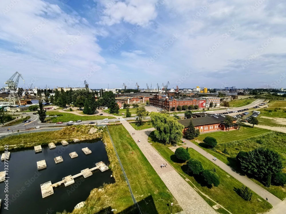 Obraz premium Aerial view of the Gdańsk Shipyard, Poland, showcasing historic industrial buildings, shipyard cranes, and a green urban landscape. A place of rich history and maritime heritage.