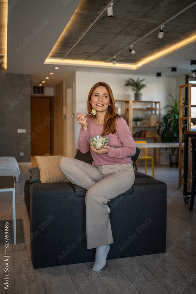 Obraz premium Smiling woman eating fresh salad on sofa at home