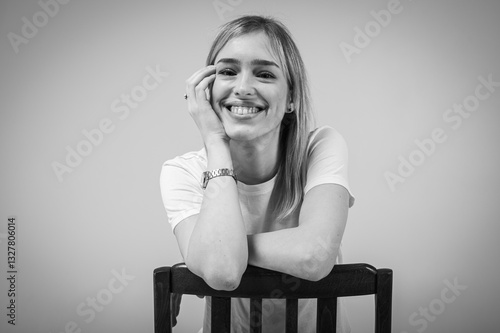 black and white portrait, happy, emotional young girl with blond hair smiling and leaning on a wooden chair on a neutral background. Cheerful woman, confidence, joy, positivity, place for inscription