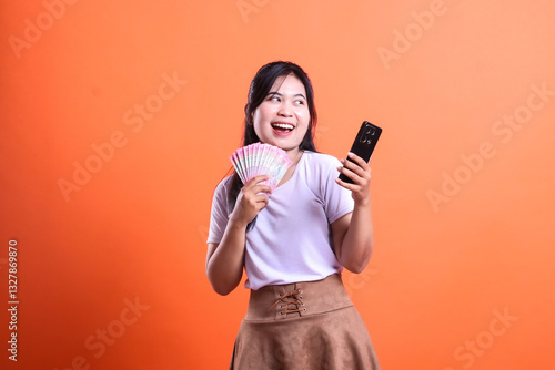 Photos Young woman holding money and phone smart phone device in orange background expr