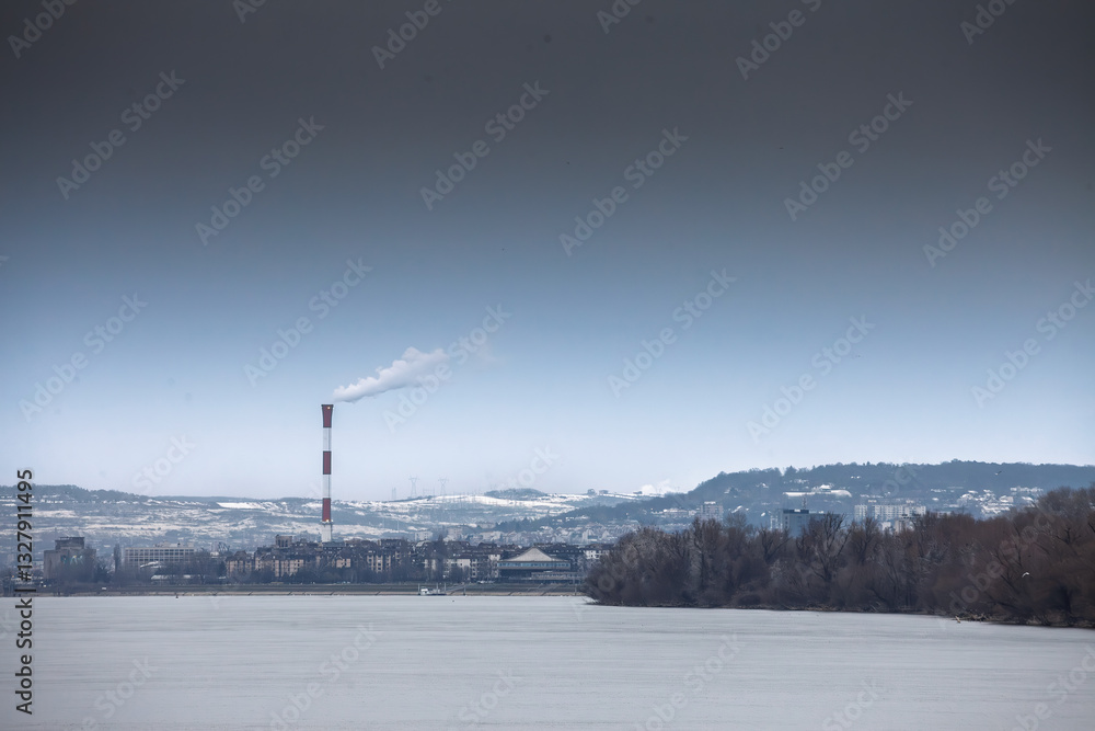 Fototapeta premium Industrial chimney emitting smoke causing visible air pollution over the frozen Danube river during wintertime in Belgrade, Serbia, illustrating environmental issues and industrial impact.