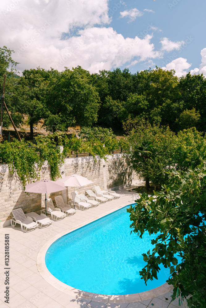 Fototapeta premium White sun loungers stand on the shore of an oval pool in a green garden