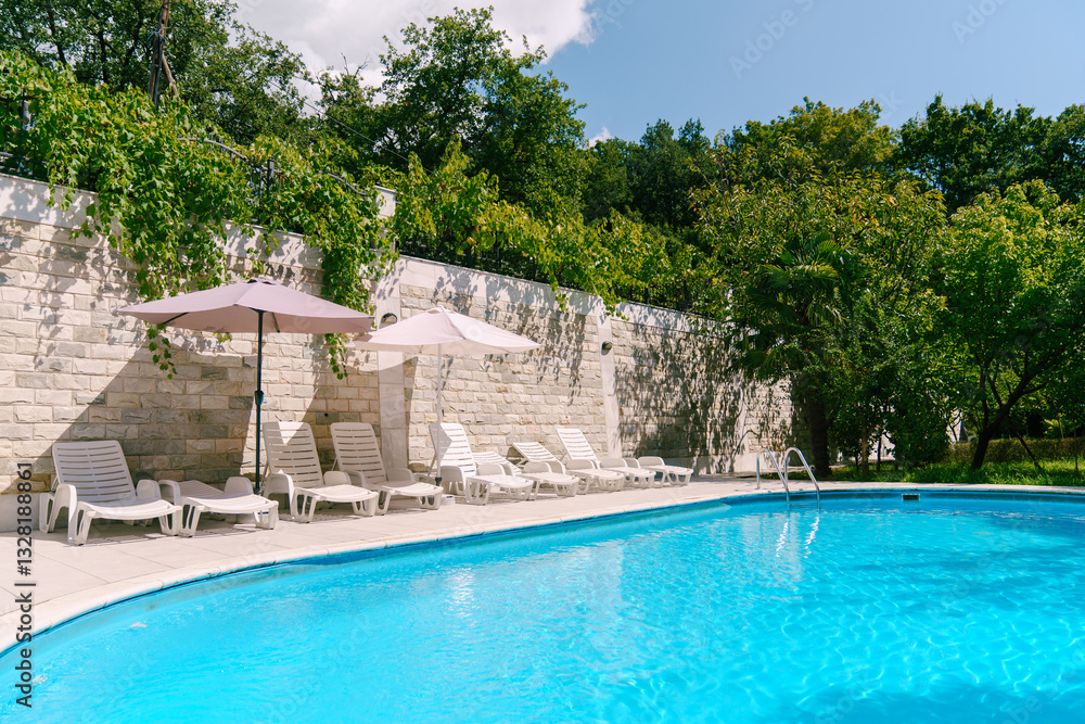 Fototapeta premium Swimming pool among green trees in the courtyard near a brick fence with sun loungers and sun umbrellas