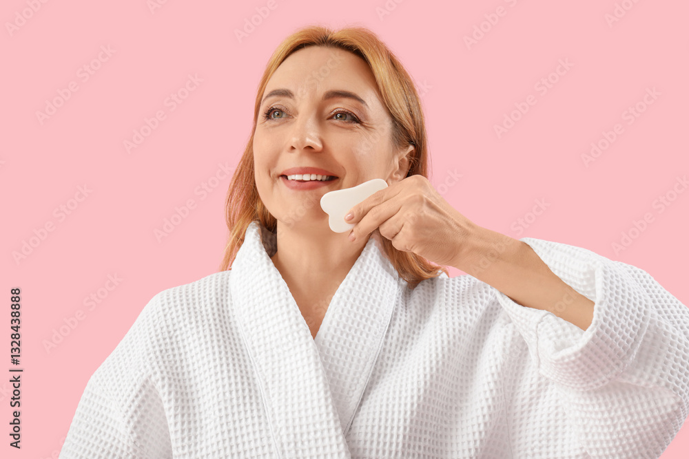 Beautiful happy mature woman in bathrobe doing face building exercise with guasha tool on pink background Beautiful happy mature woman in bathrobe doing face building exercise with guasha tool on pink background