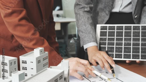 Two businesswomen sit at a desk in a meeting, discussing property development while building a model of city tower. real estate investments, zoning laws, sustainable energy solutions. net zero