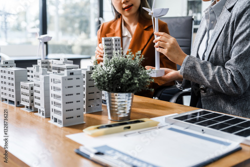 Two businesswomen sit at a desk in a meeting, discussing property development while building a model of city tower. real estate investments, zoning laws, sustainable energy solutions. net zero