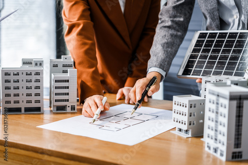 Two businesswomen sit at a desk in a meeting, discussing property development while building a model of city tower. real estate investments, zoning laws, sustainable energy solutions. net zero