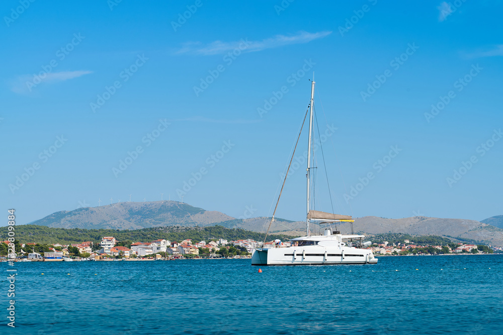 A white sailing boat rests quietly in calm waters with a picturesque coastline and mountains in the background. A white sailing boat rests quietly in calm waters with a picturesque coastline and mountains in the background.