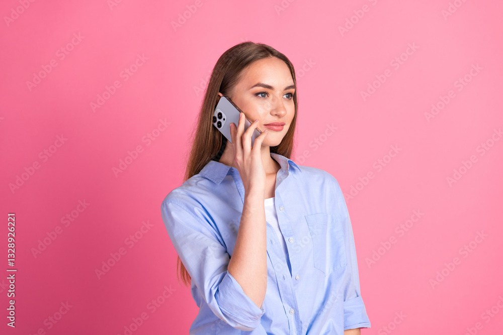Confident young woman with long brown hair talks on smartphone, wearing a casual blue shirt, standing against a vibrant pink background Confident young woman with long brown hair talks on smartphone, wearing a casual blue shirt, standing against a vibrant pink background