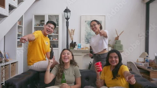 Group of Asian friends watching TV together in a cozy living room, cheering with drinks in hand, smiling and reacting with excitement while sitting on a couch during a casual home gathering