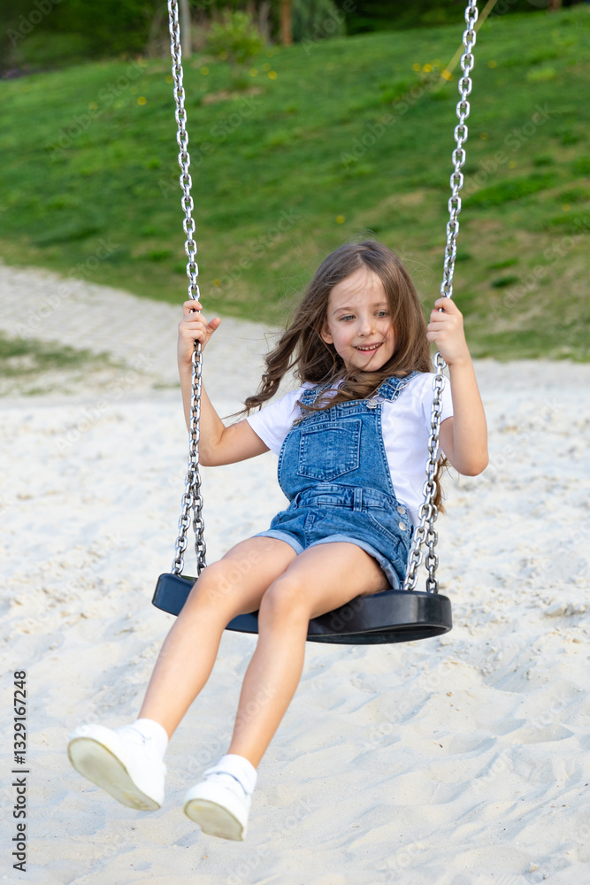 Happy joyful little girl in white summer dress rides on a swing on a sandy beach with green lawn background or on playground in children park, on a sunny day. Vacation concept. Children's Day Happy joyful little girl in white summer dress rides on a swing on a sandy beach with green lawn background or on playground in children park, on a sunny day. Vacation concept. Children's Day