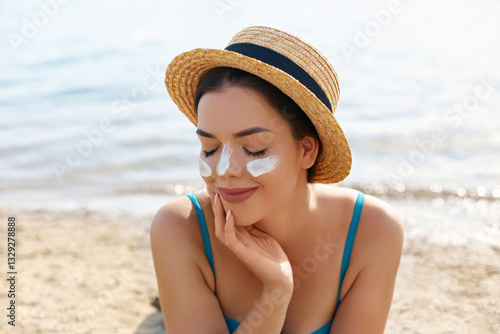 A young woman with a straw hat sits on the sandy beach, smiling with joy as she applies sun cream on her face. The sun shines brightly over the calm sea photo