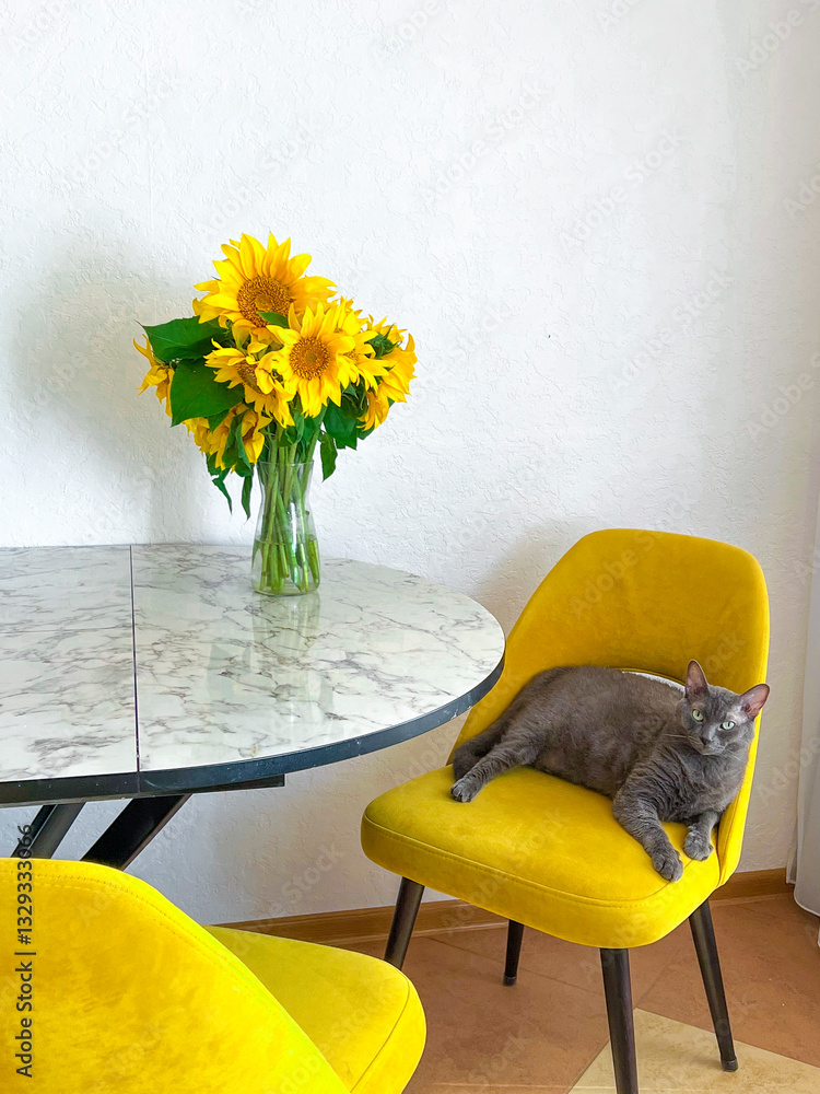 A grey cat is lying on a yellow armchair near a table with a bouquet of sunflowers