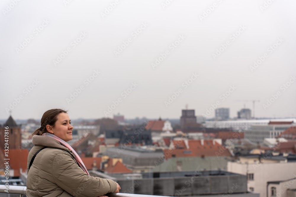 Smiling woman in warm jacket and scarf leaning on railing and enjoying panoramic city view on an overcast day. Female traveler admiring urban skyline with historic rooftops and modern buildings. Smiling woman in warm jacket and scarf leaning on railing and enjoying panoramic city view on an overcast day. Female traveler admiring urban skyline with historic rooftops and modern buildings.