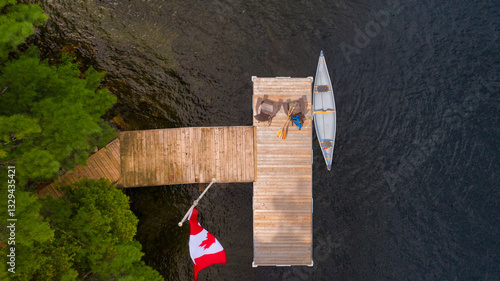 Fototapeta Naklejka Na Ścianę i Meble -  Aerial view of a wooden dock on a lake in Muskoka, Ontario. Two Adirondack chairs face the blue water, with a Canadian flag waving. A yellow canoe is tied to the dock with paddles and life jackets.
