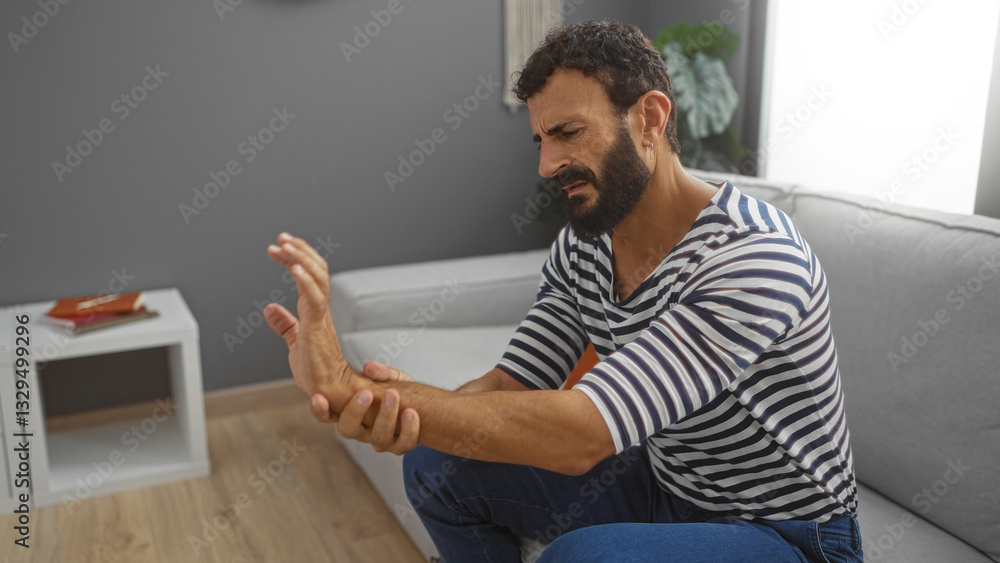 Young man with beard wearing striped shirt sitting in living room holding his painful wrist with a discomforted expression, highlighting indoor domestic setting. Young man with beard wearing striped shirt sitting in living room holding his painful wrist with a discomforted expression, highlighting indoor domestic setting.