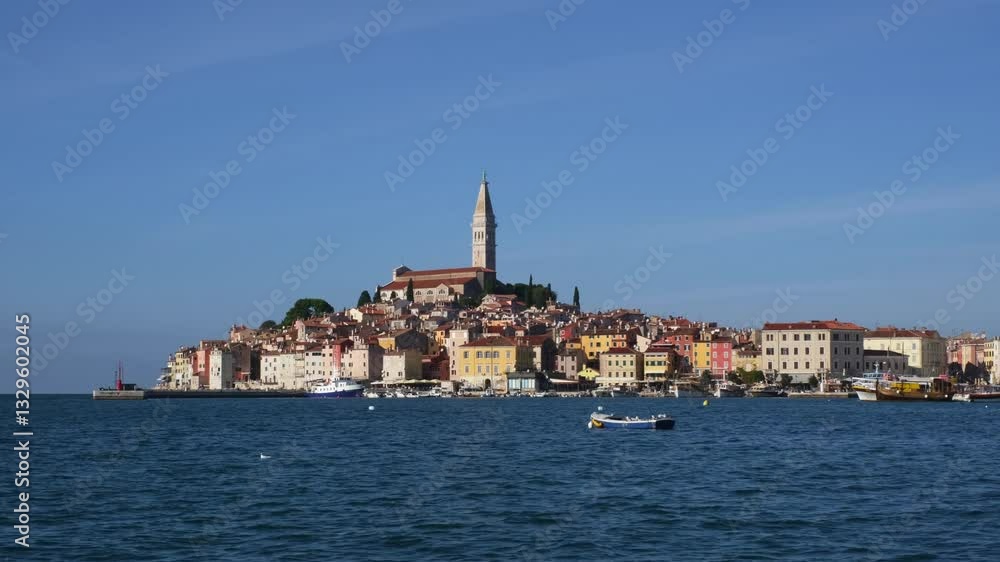 View of Rovinj, Croatia across the bay on a sunny day. The famous cathedral on the top of the hill surrounded by old medieval houses and blue wavy sea surface with a little boat in the foreground. View of Rovinj, Croatia across the bay on a sunny day. The famous cathedral on the top of the hill surrounded by old medieval houses and blue wavy sea surface with a little boat in the foreground.