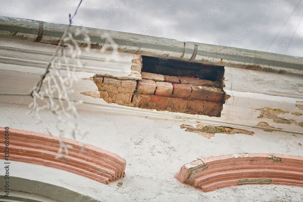 Old historic building with peeling white paint and large hole in cornice, exposing red bricks and revealing signs of decay, deterioration, and structural damage over time. Broken, damaged molding