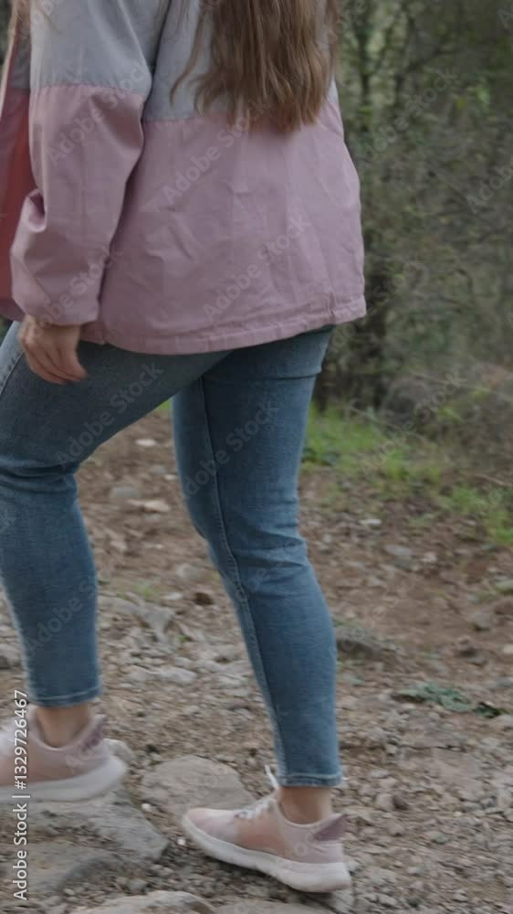 Vertical video. Woman hiking on a rugged dirt trail in a dense forest, stepping over rocks while wearing a cap, pink jacket, and jeans, surrounded by tall trees and lush vegetation Vertical video. Woman hiking on a rugged dirt trail in a dense forest, stepping over rocks while wearing a cap, pink jacket, and jeans, surrounded by tall trees and lush vegetation