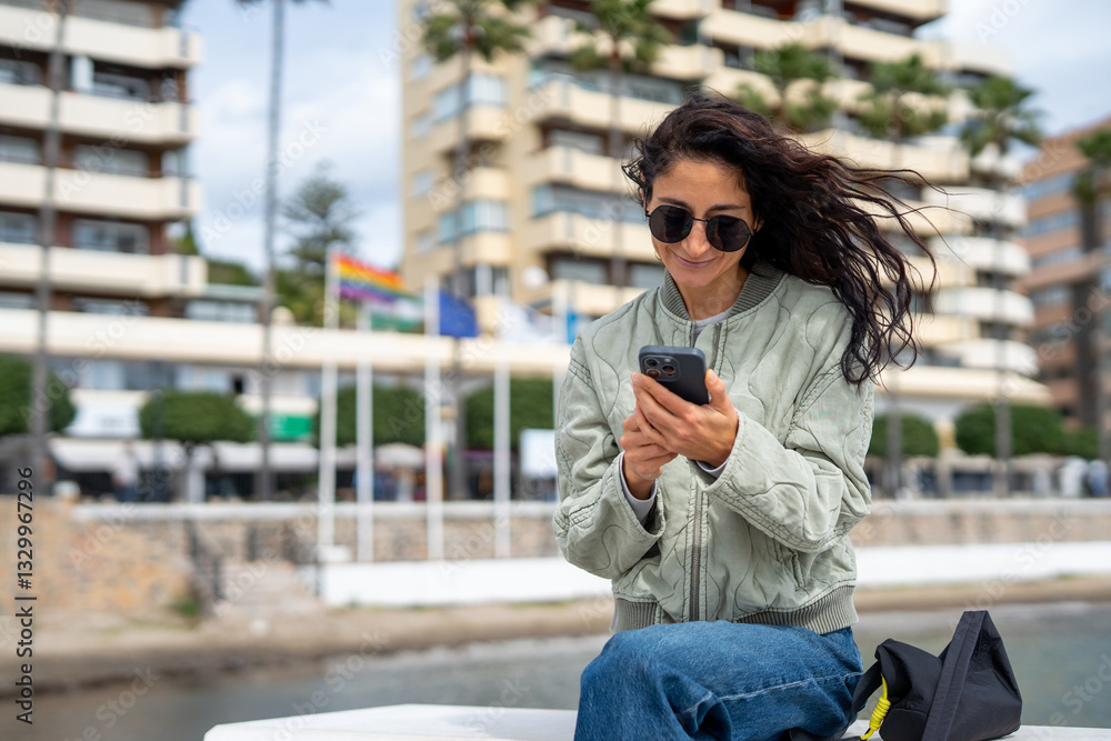 Fototapeta premium A fashionable young woman with sunglasses and a quilted jacket sits near the waterfront, smiling as she types on her smartphone, with flags and modern buildings in the background