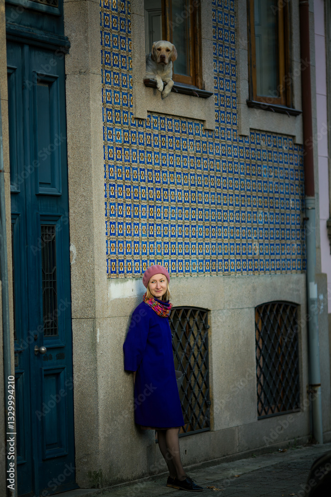 A woman in a stylish coat and beret leans against the wall of a traditional Portuguese house adorned with azulejos, while a dog peers out from the window, creating a charming and serene scene.