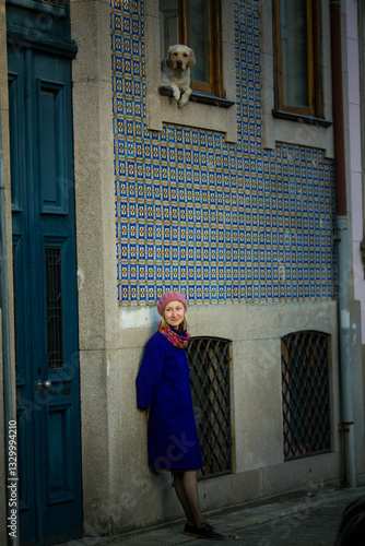 Obraz na płótnie A woman in a stylish coat and beret leans against the wall of a traditional Portuguese house adorned with azulejos, while a dog peers out from the window, creating a charming and serene scene. w salonie