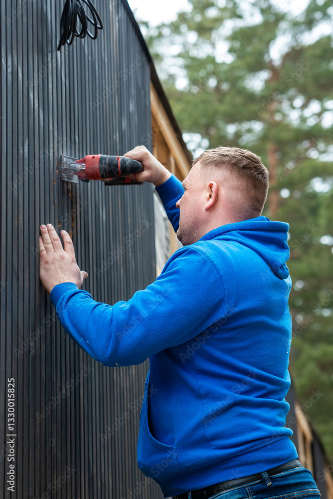 Naklejka premium A man in a blue hoodie operates a power tool on a dark vertical paneled wall of a house under construction, surrounded by tall trees and materials.