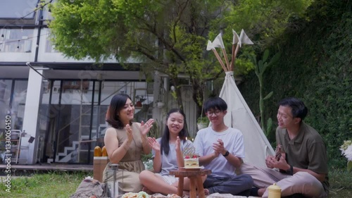Asian family and friends celebrating a birthday picnic outdoors with a cake, clapping and smiling near a canvas tent, surrounded by food, greenery, and cozy backyard decorations