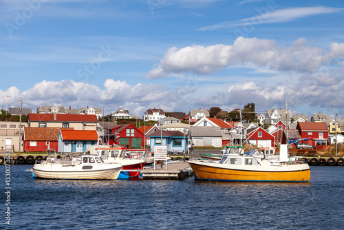 A scenic view of Borhaug, Lista in Norway, features a tranquil harbor with blue water and neatly arranged boats. Traditional red and white wooden houses line the shore under a cotton-cloud sky.