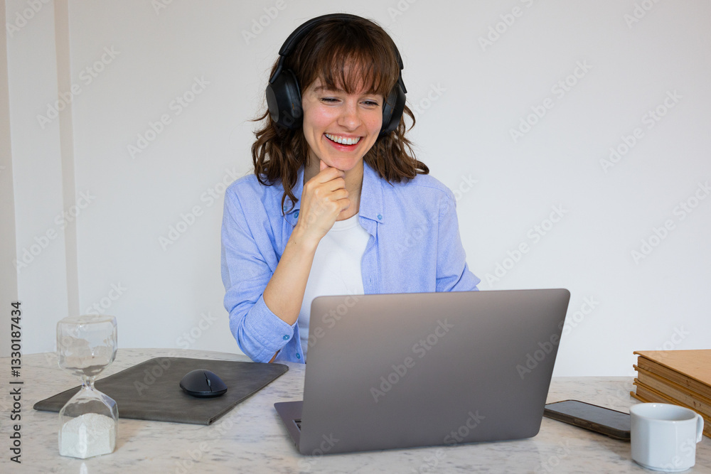 Beautiful intelligent smiley curly brunette in headphones sitting at the table with the laptop, attending the online business meeting and listening attentively to her colleagues Beautiful intelligent smiley curly brunette in headphones sitting at the table with the laptop, attending the online business meeting and listening attentively to her colleagues