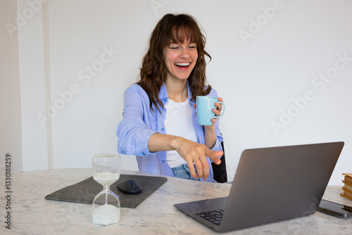 Beautiful smiley curly brunette sitting at the table with the laptop and the cup of coffee, laughing and watching videos and netflix