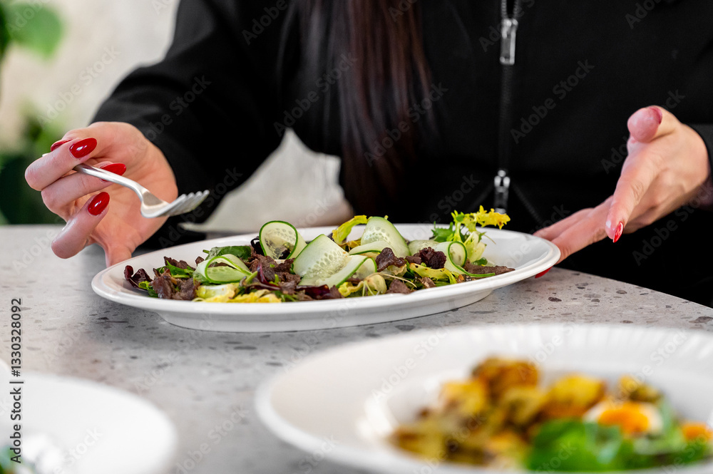 A white plate with a fresh salad of cucumber slices, mixed greens, and yellow flowers. A hand with red-painted nails holds a fork, set on a light table. A white plate with a fresh salad of cucumber slices, mixed greens, and yellow flowers. A hand with red-painted nails holds a fork, set on a light table.