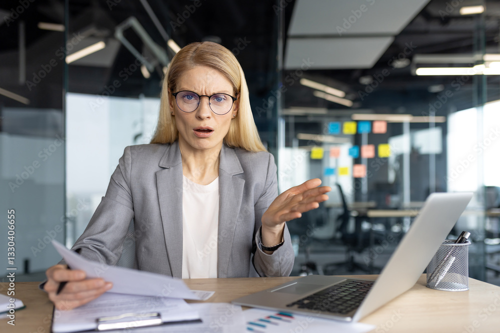 An office worker looks shocked and displeased with the documents she is reviewing while working at her desk with a laptop. An office worker looks shocked and displeased with the documents she is reviewing while working at her desk with a laptop.