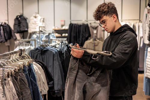 Fotografie Teenage boy choosing clothes in retail store, young man with glasses and curly hair shopping independently, adolescent exploring fashion options
