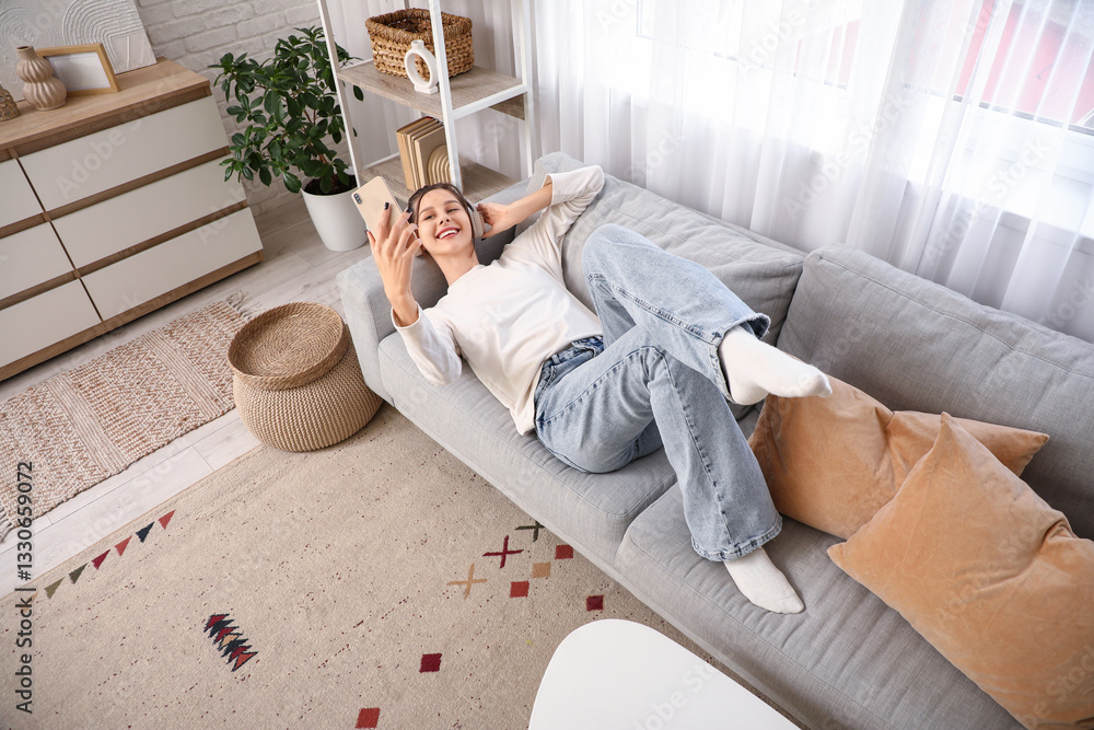Fototapeta premium Young woman in headphones taking selfie on sofa in living room