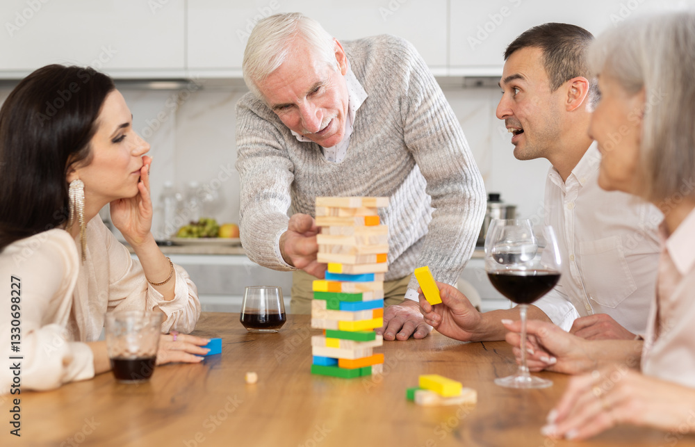 Mature parents and son with girlfriend are having fun, playing building tower with wooden blocks game. Participant is removing wooden block bricks from tower and others are watching. Mature parents and son with girlfriend are having fun, playing building tower with wooden blocks game. Participant is removing wooden block bricks from tower and others are watching.