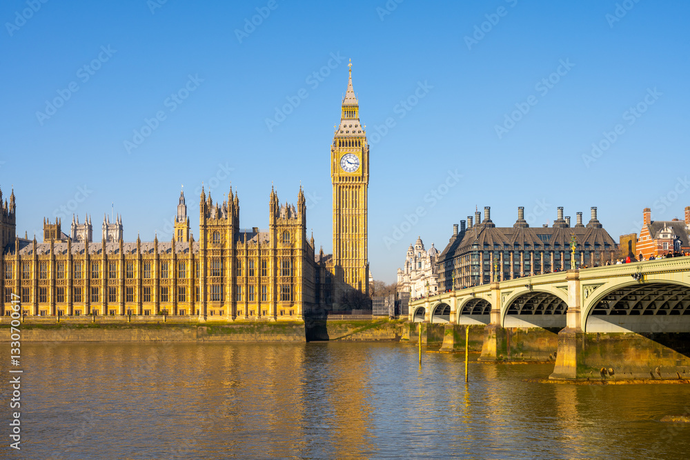 Fototapeta premium Sunlight reflects on the River Thames as the Houses of Parliament and Big Ben stand proudly in London. Westminster Bridge connects the iconic landmarks, depicting a vibrant day.