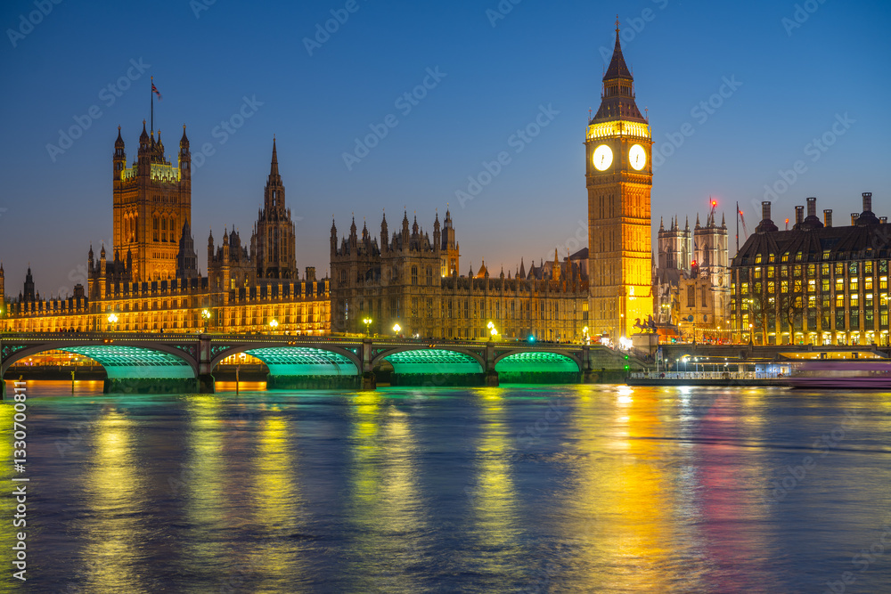 Fototapeta premium Beautiful lights illuminate the Houses of Parliament and Big Ben at night in London, with Westminster Bridge reflecting in the river. The historic architecture is striking against the twilight sky.