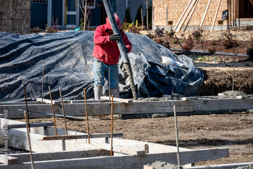 Construction worker in red sweatshirt, jeans, and rubber boots holding pumper hose pouring fresh cement into wood foundation forms, new housing development
