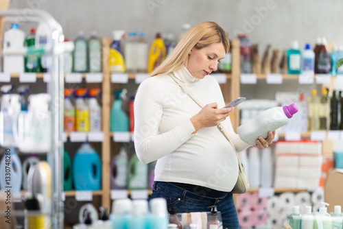Thoughtful pregnant female shopper scanning barcode on bottle of fabric softener using phone to make mobile payment in home care products shop. Concept of technology enhancing shopping experience..