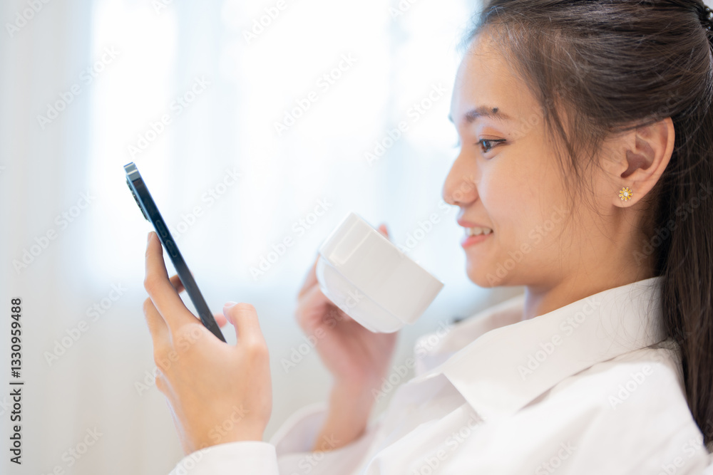 Close-up of Asian woman smiling and looking at her smartphone in hotel bedroom. She is eager to relax on her vacation and enjoy a delicious drink before heading out on a night of adventure.