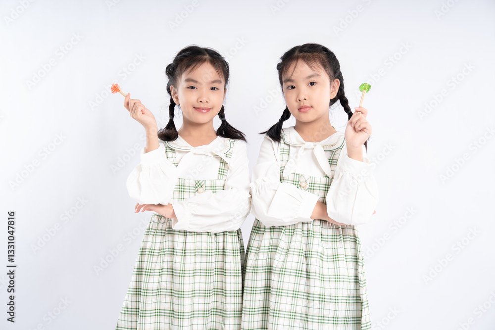 Twin girls posing with lollipops against a plain backdrop in matching green plaid dresses with white tops. Twin girls posing with lollipops against a plain backdrop in matching green plaid dresses with white tops.