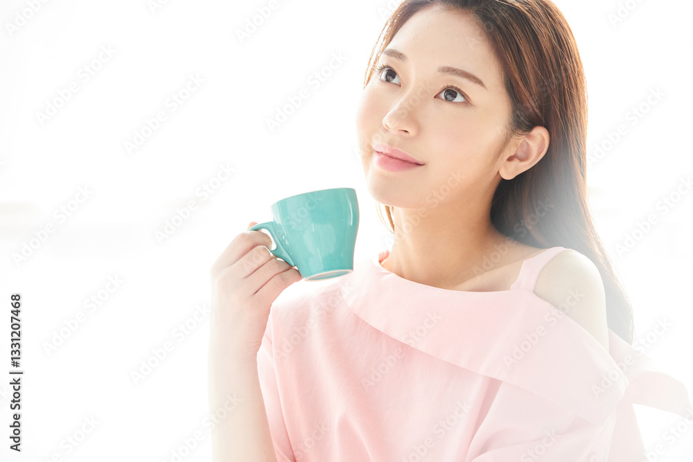 A beautiful young woman is drinking coffee from a blue cup, standing in front of a sunlit window with her back to the sunlight A beautiful young woman is drinking coffee from a blue cup, standing in front of a sunlit window with her back to the sunlight