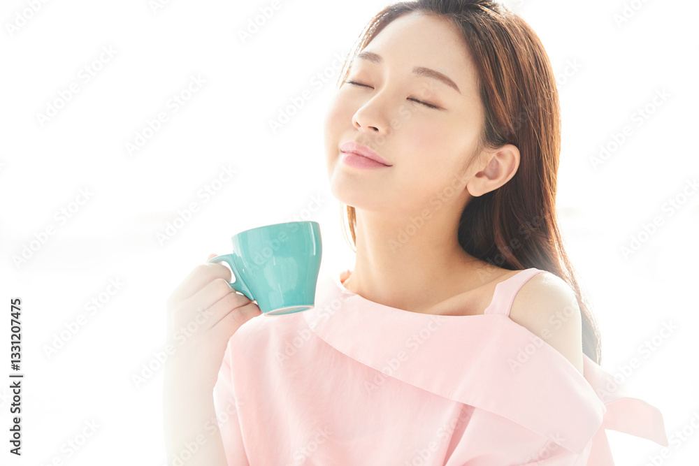 A beautiful young woman is drinking coffee from a blue cup, standing in front of a sunlit window with her back to the sunlight A beautiful young woman is drinking coffee from a blue cup, standing in front of a sunlit window with her back to the sunlight